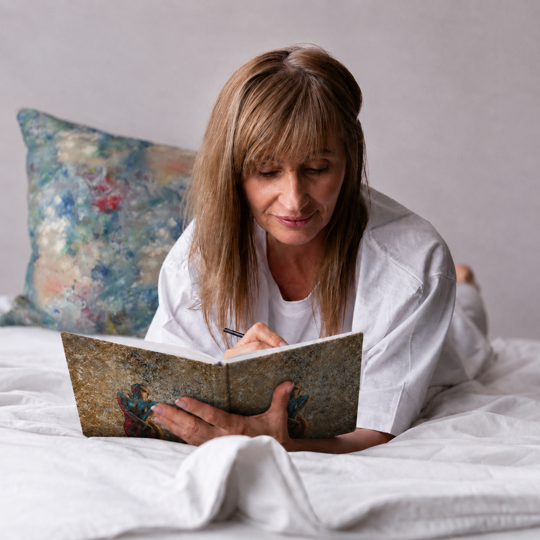 Woman writing in a Charmed Life HOME Eternal Glow journal beside a Red Snapper print pillow on white bedding.